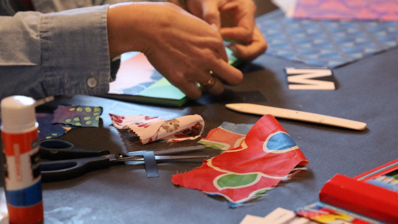 person out of frame working on artwork, seated at table with a variety of art materials
