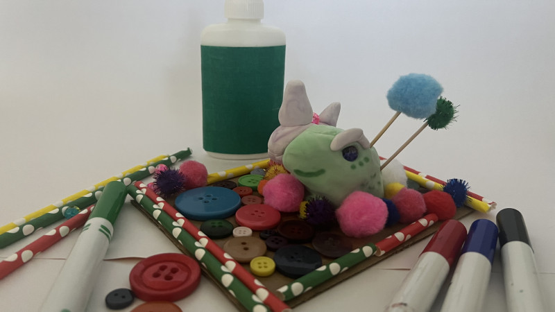Image of an arts and craft project. The craft is a model magic sculpture, Hal is a lizard, and half is a unicorn. It sits on a cardboard plate with decorative buttons and pompoms. surrounded by art supplies, inclusive of markers, buttons, and glue.
