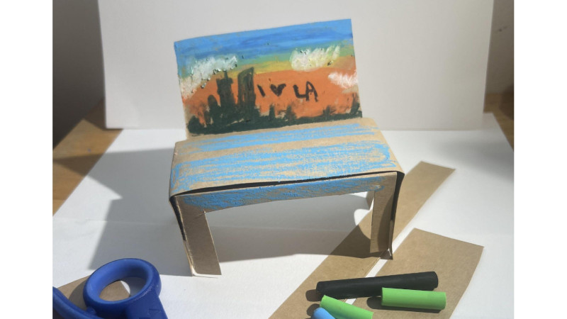 The image is of a 3D paper bench with an oil pastel drawing on the paper sculpture. The drawing on the paper bench is of the LA landscape and says " I love LA".