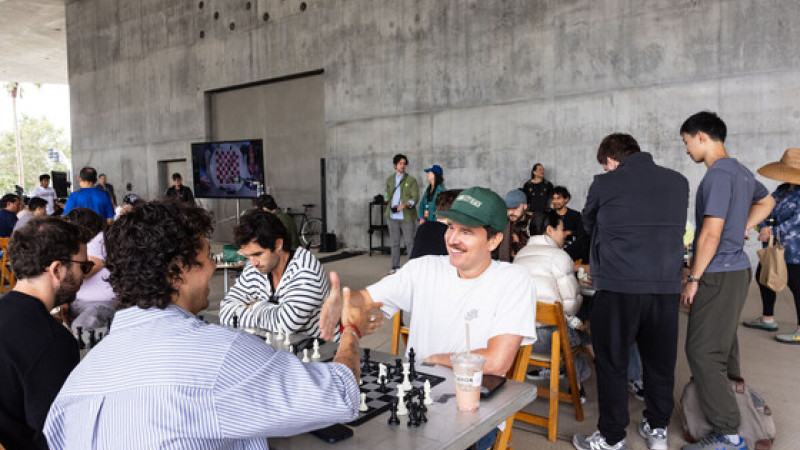 Photograph of chess players, two are shaking hands over a chess board and smiling