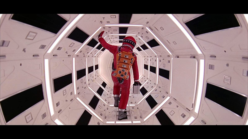 Image of a figure in a red space suit walking through an octagonal hallway lit and interspersed with bars of lights 
