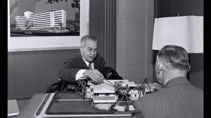 Man at desk pointing at an architectural model of a low building with palm trees, another man seated across from him, framed rendering of a high-rise hospital on the wall behind.