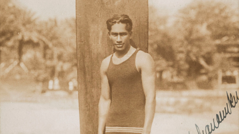A sepia toned photograph showing a figure with short dark hair in a bathing costume standing in front of a surfboard that reads “DUKE” on the beach, handwritten lettering in the bottom right corner reads “Duke P. Kahanamoku” above the word “Waikiki” and “1917”