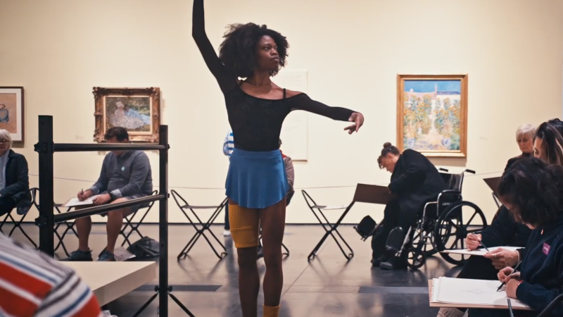 A woman stands in a dancers stance in front of people sitting and drawing on paper