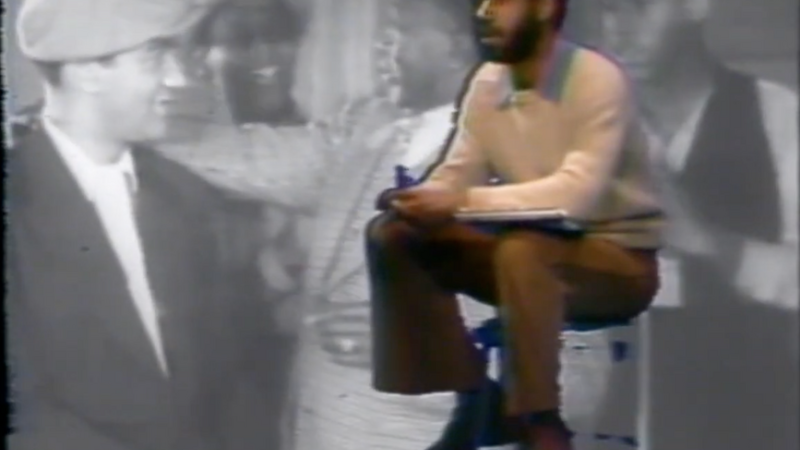A color image of a Black male figure with a beard and short hair dressed in a cream sweater and brown pants sitting on a stool placed on top of black and white historical footage of various figures in the background