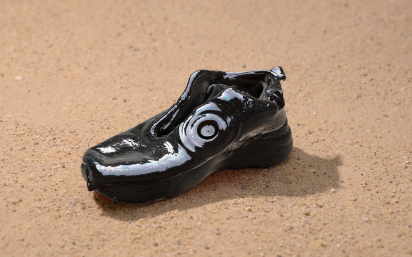 Glossy black shoe with circular form on the side, with thick sole, sitting on sand.