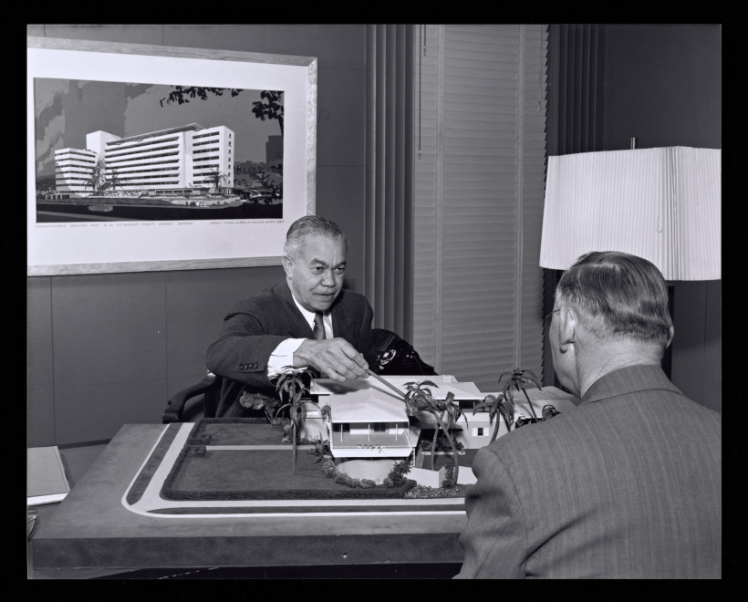 Man at desk pointing at an architectural model of a low building with palm trees, another man seated across from him, framed rendering of a high-rise hospital on the wall behind.