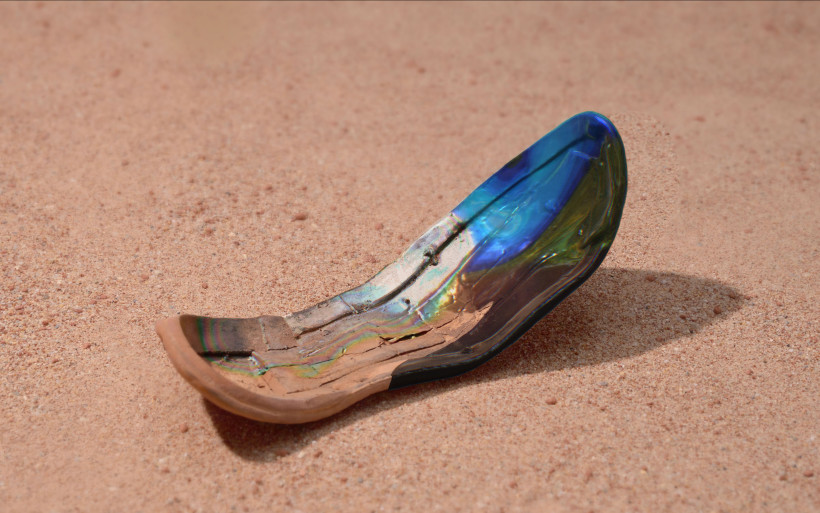 Fragment of a shoe with iridescent blue and gold patina resting on reddish desert sand, showing weathered layers and surface cracking.