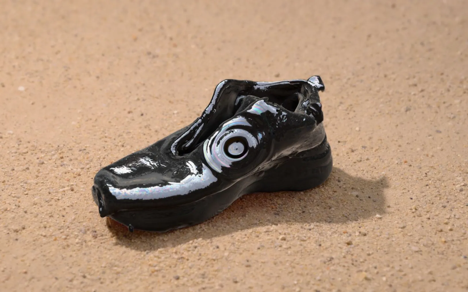 Glossy black shoe with circular form on the side, with thick sole, sitting on sand.