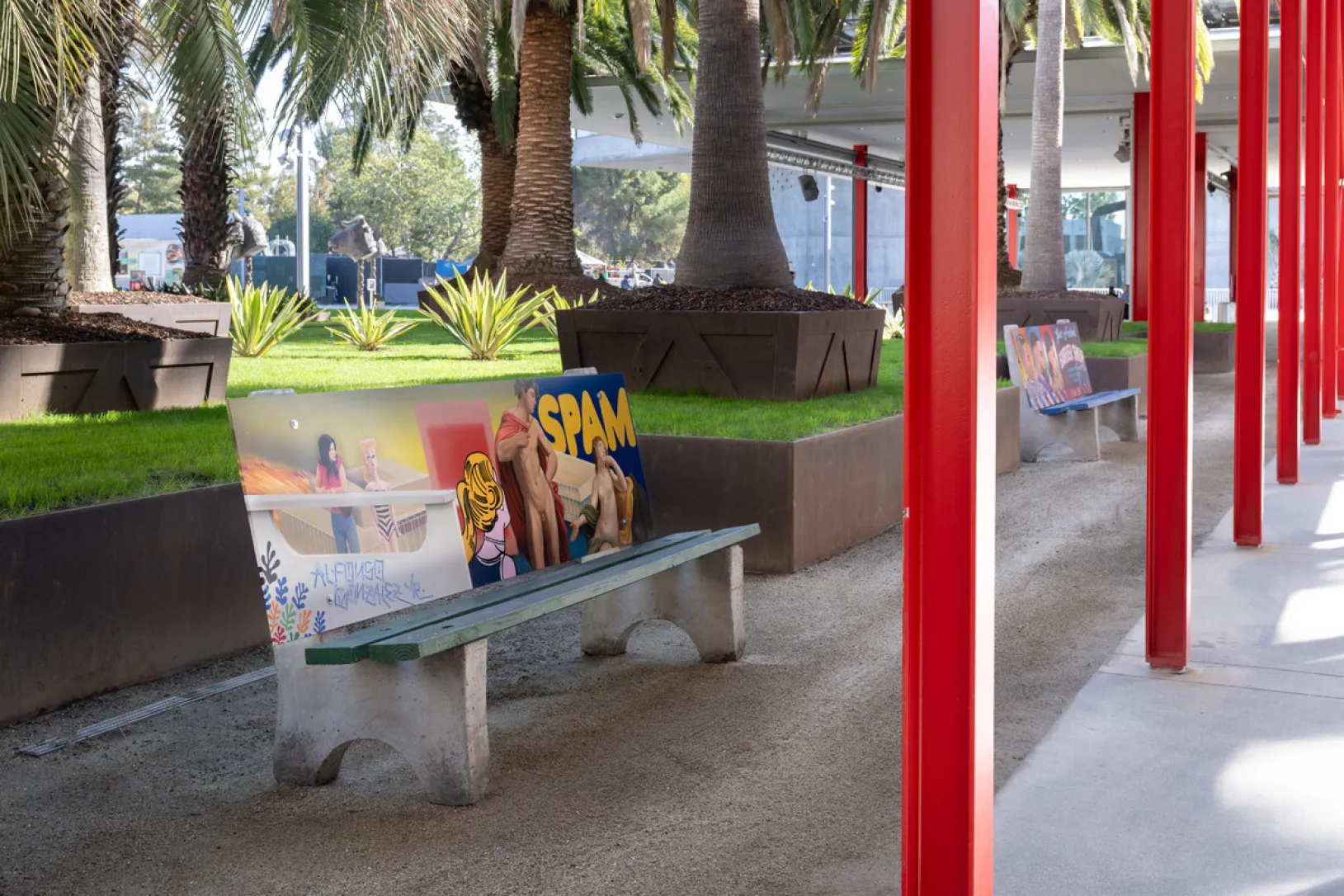 Painted panels leaning on concrete benches along a museum pathway, one reading "SPAM" with figures, red columns and palm trees in the background.