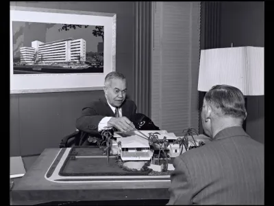 Man at desk pointing at an architectural model of a low building with palm trees, another man seated across from him, framed rendering of a high-rise hospital on the wall behind.